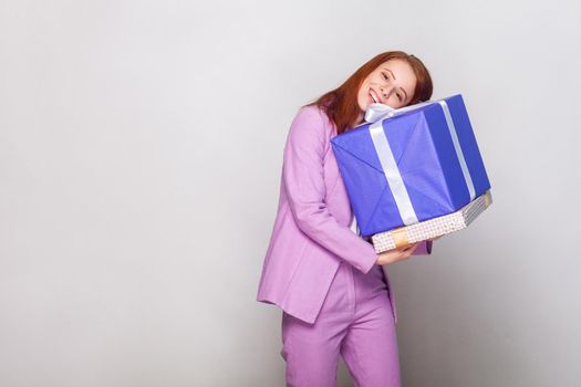 Happy birthday concept! Foxy cute girl hugging her gifts and looking at camera with toothy smile. Studio shot, gray background