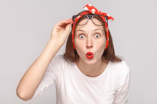 are you serious? portrait of wondered surprised young woman in white t-shirt with freckles, glasses, red lips and head band looking at camera with big eyes. indoor shot, isolated on gray background.