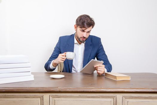 Technology, people and business concept - handsome man with beard and brown hair and blue suit and tablet pc computer and some books and cup drinking coffee or tea sitting in the office. .Isolated on white background. .