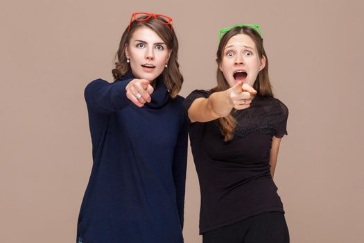 Beautiful women standing near each other and pointing fingers at camera and shock. Studio shot, light brown background