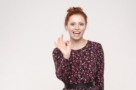 Confident woman with charming smile making ok gesture being agreeable. Cute smiling female with red hair showing okay sign. Studio shot on gray background