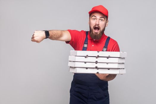 Hurry up! time is gone. Young amazed delivery man with beard in blue uniform and red t-shirt, holding stack of cardboard pizza boxes on grey background. Indoor, studio shot, isolated, copy space.