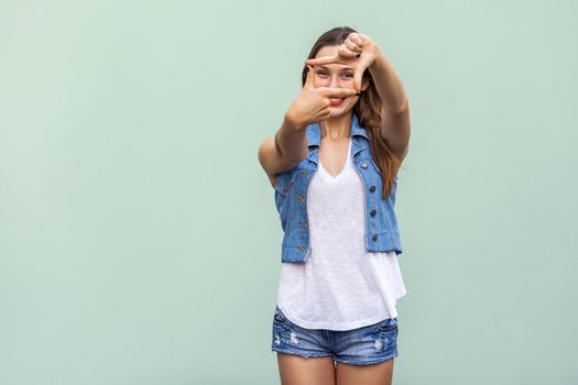 Creative young adult woman with freckles, making a frame gesture with her fingers as she looks through to visualise a project or the composition of a photograph. Indoor studio shot on light green background