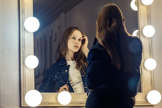 Young beautiful sexy woman looking in a mirror, touching her hair sitting on chair at dressing room with vintage mirror dark room. Studio shot