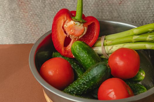 Vegetables lie in a metal bowl: tomatoes, asparagus, cucumbers, red bell peppers . on a wooden board and brown background. back gray background. place for text.