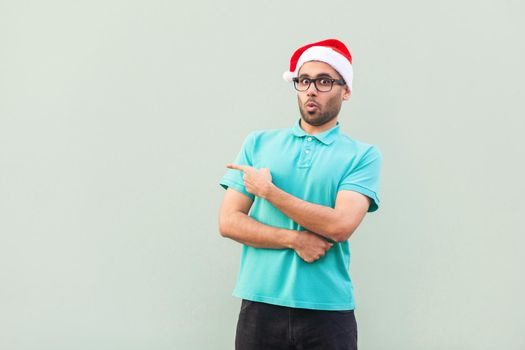 Amazed bearded man in christmas cup with beard in shoked. Pointing away while standing isolated on gray background. Looking at camera with big eyes and open mouth. Studio shot