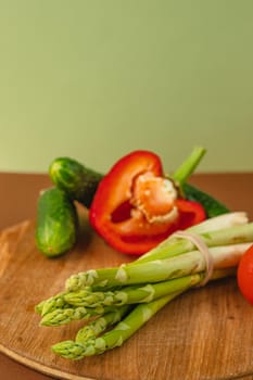 Vegetables lie on a wooden board: tomatoes, asparagus, cucumbers, red bell peppers. brown, light green background. place for text