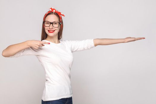 happy toothy smiley portrait of beautiful emotional young woman in white t-shirt with glasses, red lips and head band pointing and showing copy space. indoor studio shot, isolated on gray background.