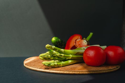 Vegetables lie on a wooden board: tomatoes, asparagus, cucumbers, red bell peppers. brown, dark gray background. place for text