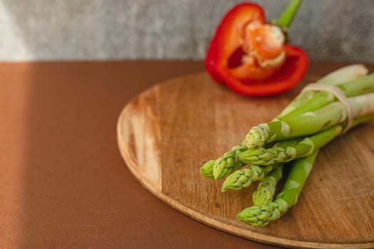 branches of fresh green asparagus on a wooden board, red big pepper on the background. brown background, top view. Basic trend concept with copy space
