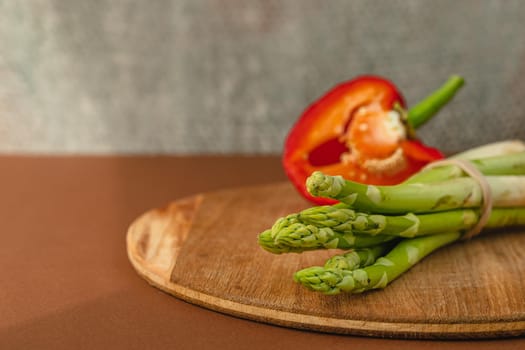 branches of fresh green asparagus on a wooden board, red big pepper on the background. brown background. Basic trend concept with copy space