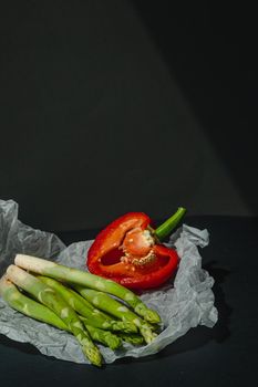 fresh green asparagus and half of red sweet, large pepper lie on white parchment on a dark gray background. proper nutrition. Lifestyle. place for text