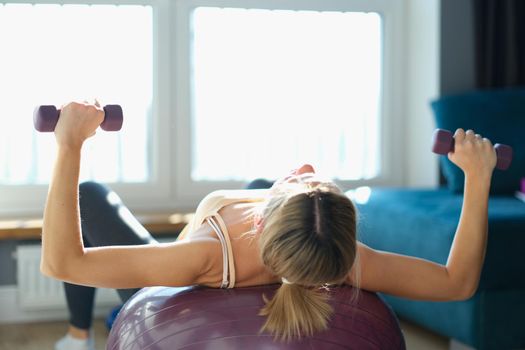 Young woman doing bench press with dumbbells and stabilizing ball at home. Home fitness workout concept