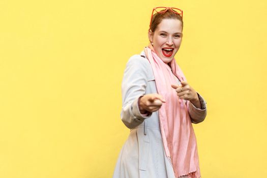 Happy cheerful girl with freckles, wearing coat, pink scarf, red glasses, looking and pointing fingers at camera . Studio shot, yellow background