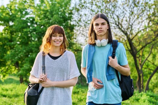 Portrait of teenage young guy and girl students looking at camera, outdoor. Couple of young people with backpacks, on green lawn of campus. Teenagers, lifestyle, joy, friendship, education concept