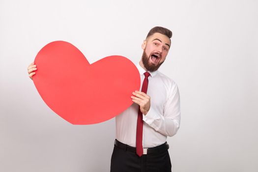Funny businessman toothy smiling, holding big red heart. Indoor, studio shot, isolated on gray background