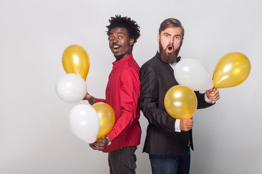 Two shocked friend wearing red shirt and black jacket holding many air balloon and looking at camera with shocked face. Indoor shot