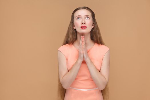 Oh God please forgive me. portrait of emotional cute, beautiful woman with makeup and long hair in pink dress. indoor, studio shot, isolated on light brown or beige background.