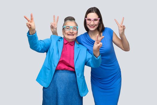 Grandmother with Granddaughter showing peace or victory sign, looking at camera with toothy smile. Relations in the family. happiness and family relationship. Studio shot, isolated on gray background