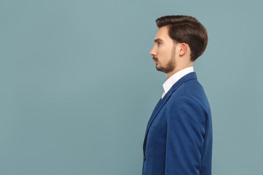 side view or profile view of serious handsome bearded businessman in blue suit and white shirt. Indoor studio shot, isolated on light blue background