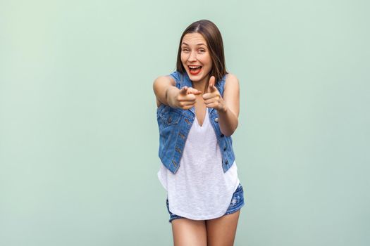 Happy cheerful teenage girl with freckles, casual style clothes, looking and pointing fingers at camera indoors. Studio shot, light green background