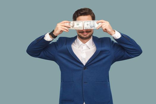 Handsome happy businessman in blue jacket and white shirt standing and holding and covered many dollars in front of eyes and smiling . Indoor, studio shot isolated on light blue background.