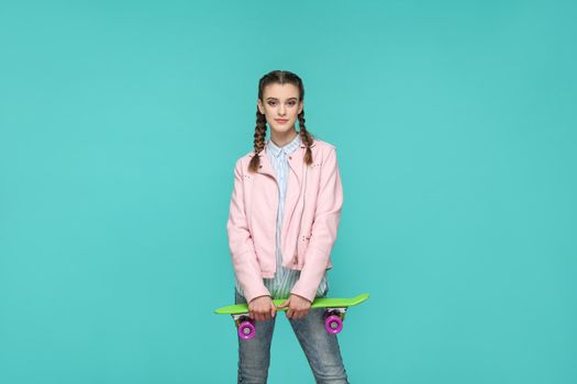 hipster beautiful girl in casual style, pigtail hairstyle and pink jacket, standing and holding skateboard and looking at camera and smiling, Indoor isolated, studio shot on blue or green background