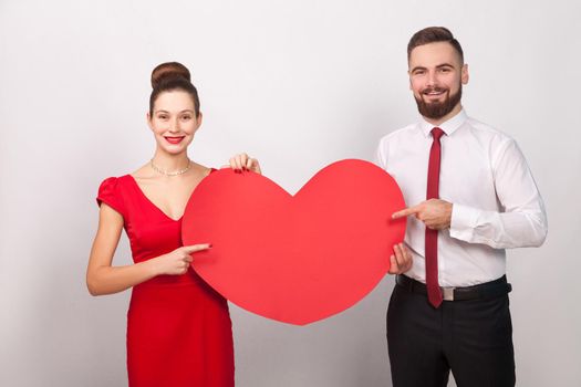 Handsome lively couple pointing finger at big heart, toothy smiling. Indoor, studio shot, isolated on gray background