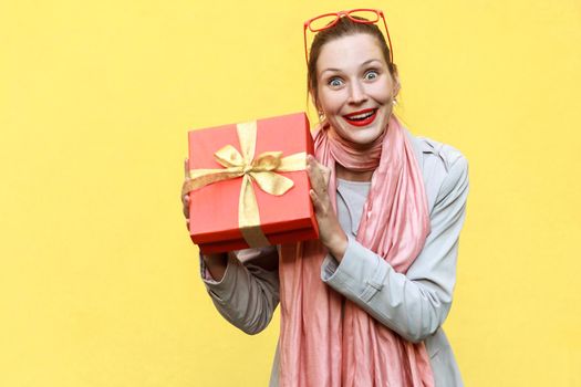 Young adult woman holding gift box and looking at camera and toothy smile. On yellow background. Studio shot