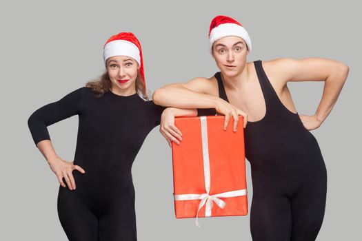 funny couple in black dress and red christmas hat holding gift box together and looking at camera. Indoor studio shot, isolated on gray background