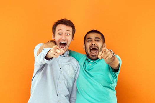 Closeup. Two happiness friends hugging each other, pointing finger and looking at camera and scream or shout. Studio shot on orange background