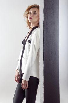 Fashionable young model posing near black wall undressing her white jacket and closed eyes. Indoor shot, gray wall
