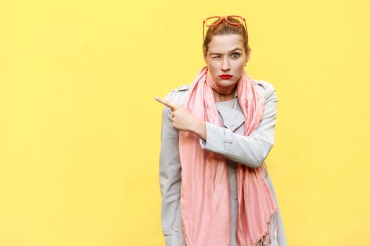 Young adult woman pointing copy space and wink. Seriously looking at camera and pointing away while standing isolated on yellow background. Studio shot