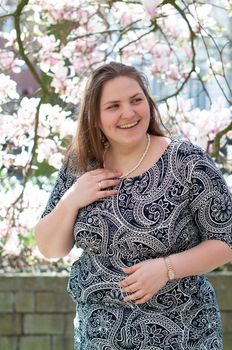 A plump, confident woman stands under a blooming magnolia and smiles. young millennial woman with brown curly hair smiling and waiting for a friend for a date. A full girl enjoys flowering and spring.