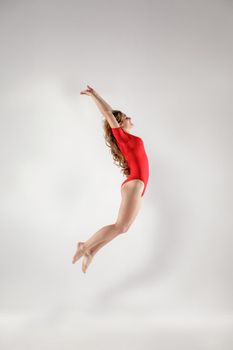 Happy beautiful blonde gymnast with curly long hair in red leotard jumping. indoor studio shot on light gray background. healthy lifestyle and leisure activity concept.