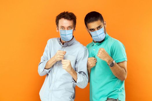 Portrait of two serious young worker men with surgical medical mask standing with boxing fists and ready to attack or defence against virus or problem. indoor studio shot isolated on orange background