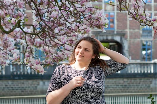 A plump, confident woman stands under a blooming magnolia and smiles. young millennial woman with brown curly hair smiling and waiting for a friend for a date. A full girl enjoys flowering and spring.