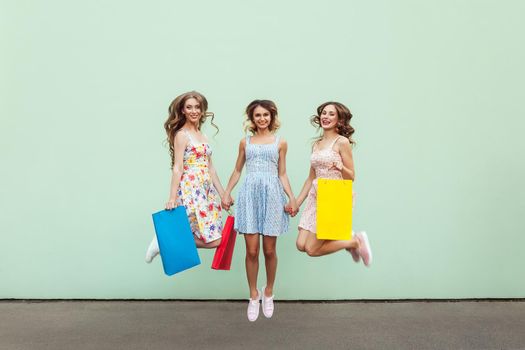 Happiness beautiful three friends jumping from happy with colorful packets after shopping. Outdoor. Green wall