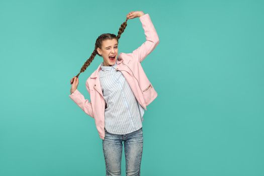 portrait of happy beautiful funny girl with blue striped t-shirt and pink jacket standing looking at camera, holding her pigtail hair and winking. indoor studio shot isolated on green background.