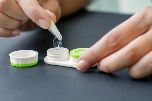 Woman takes contact lens out of container with tweezers. Black background, hands close-up, side view