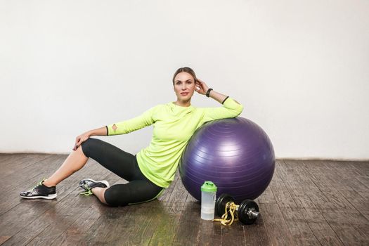 Slim beautiful woman in tight sportswear sitting on floor with big rubber gym ball, tonic drink in bottle next to dumbbell. Health care, sports activity and workout at home. indoor studio shot