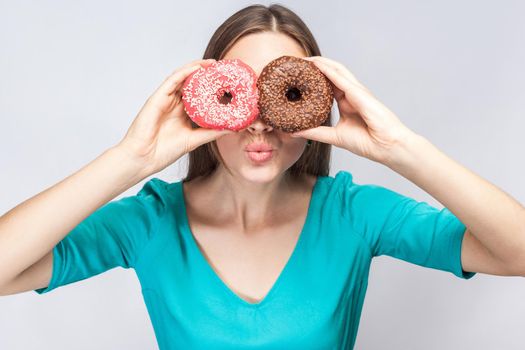 Positive young beautiful girl in blue blouse standing, holding, covering eyes with pink and chocolate donuts,looking through donuts like making glasses on grey background. Indoor, isolated, copy space