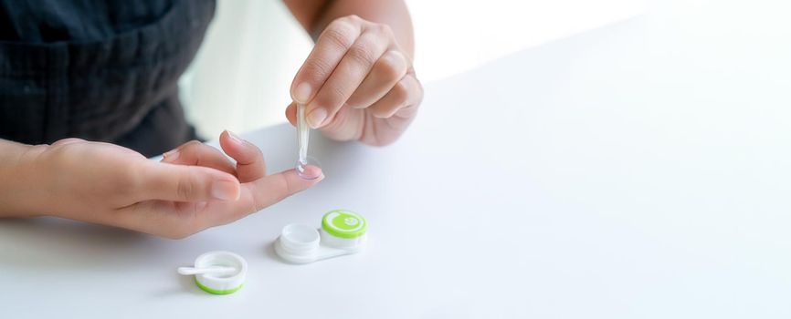 Girl puts contact lens on her index finger with tweezers. There is container for lenses nearby. Close-up, side view, banner, copy space