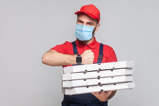 In time delivery on quarantine! man with surgical medical mask in uniform and red t-shirt standing, holding stack of cardboard pizza boxes on grey background. Indoor, studio shot, isolated, copy space