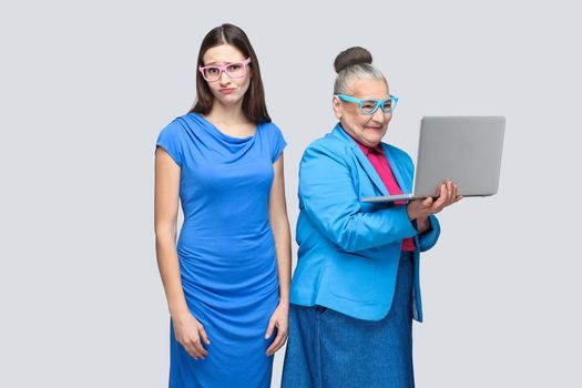 Unhappy young woman standing near happy older woman work computer . Granddaughter or daughter with grandmother. Friendship and mutual understanding. indoor, studio shot, isolated on gray background