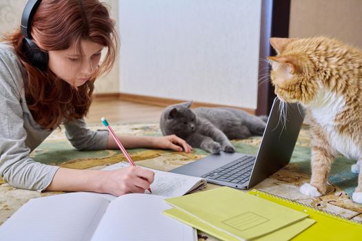 Preteen girl studying in headphones with laptop lying on floor with cats. Young female student using laptop, writing in notebook. School, learning, distance education, homework, pets lifestyle concept