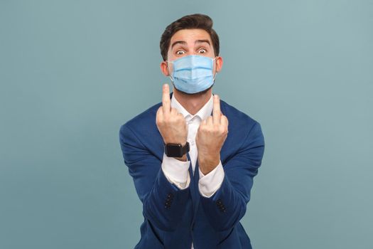 Go away from here. Young angry man with surgical medical mask showing fuck sign at camera. Business people medicine and health care concept. Indoor, studio shot on light blue background