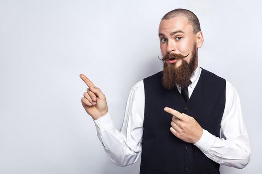 Handsome businessman with beard and handlebar mustache looking at camera, surprised and showing copy space with fingers. studio shot, on gray background.