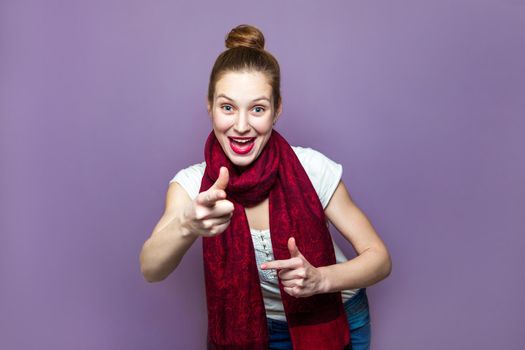 Young emotional girl with collected hair, freckles and red scarf looking excited on purple background, pointing her finger with happy funny face, looking and pointing at camera.