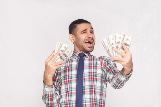 Happy joyful handsome young adult businessman in colorful checkered shirt with blue tie standing, holding fan of dollars and winking with open mouth. Indoor, studio shot, isolated on grey background